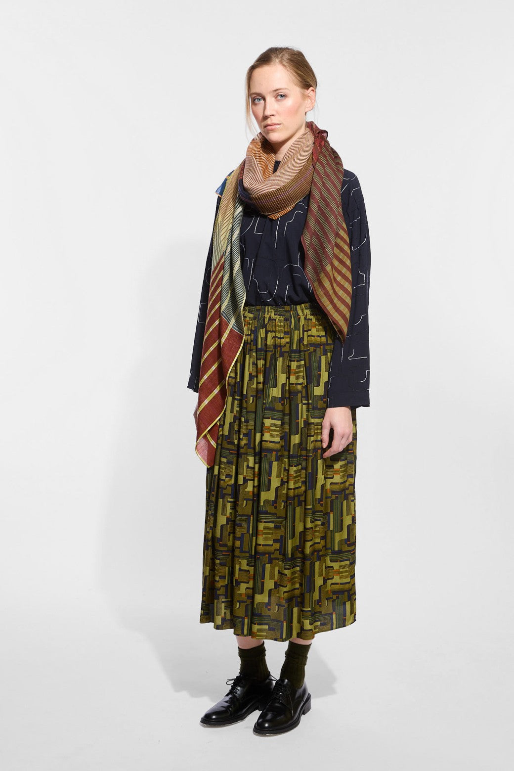 A model wearing a dark navy long-sleeve top, a pleated olive green and black patterned midi skirt, a brown scarf, and black leather shoes.