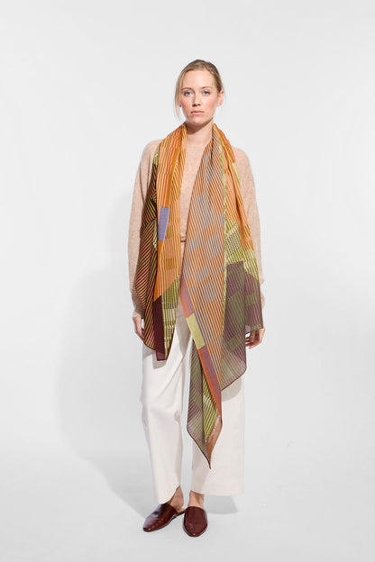 A woman wearing a beige cardigan, white wide-leg pants, brown loafers, and a long patterned scarf in orange, brown, olive green, and beige tones.