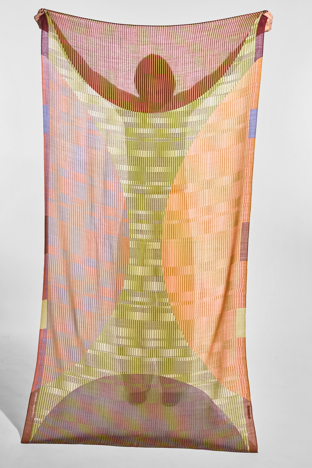 A sheer rectangular scarf or fabric panel with an abstract geometric print, hanging against a plain light gray studio background. The design features a central vertical striped motif in muted yellow-green tones, flanked by peach, orange, pink, and lavender curved and block patterns.