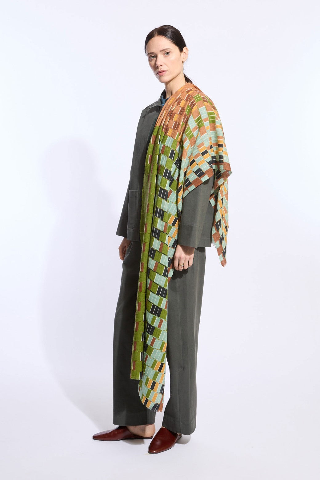 A woman models a dark olive green two-piece outfit with wide-leg trousers and a draped multicolor geometric patterned shawl over her shoulders, paired with dark brown pointed-toe flats, against a plain white studio background.