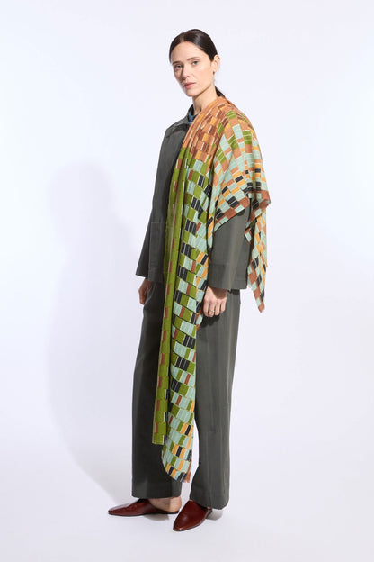 A woman models a dark olive green two-piece outfit with wide-leg trousers and a draped multicolor geometric patterned shawl over her shoulders, paired with dark brown pointed-toe flats, against a plain white studio background.