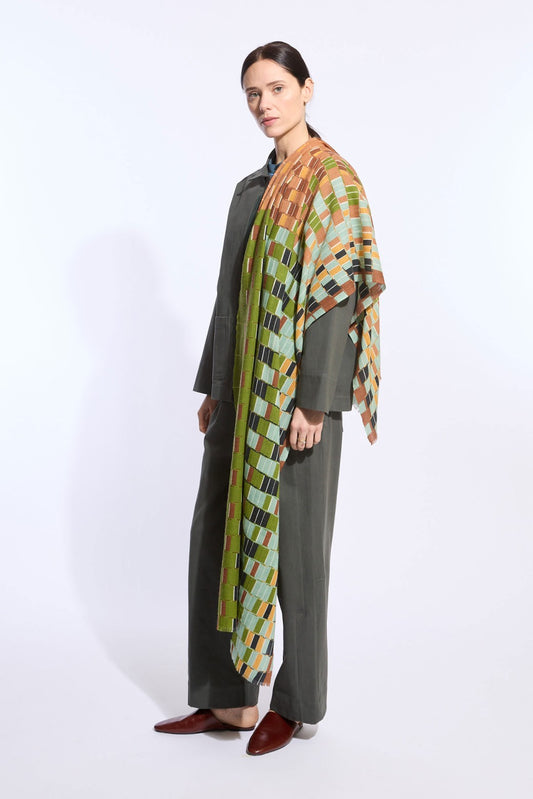 A woman models a dark olive green two-piece outfit with wide-leg trousers and a draped multicolor geometric patterned shawl over her shoulders, paired with dark brown pointed-toe flats, against a plain white studio background.