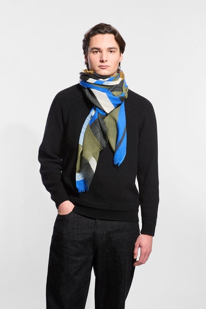 A man wearing a black knit sweater and dark jeans, styled with a patterned scarf in blue, olive green, beige, and black, against a plain light gray background.