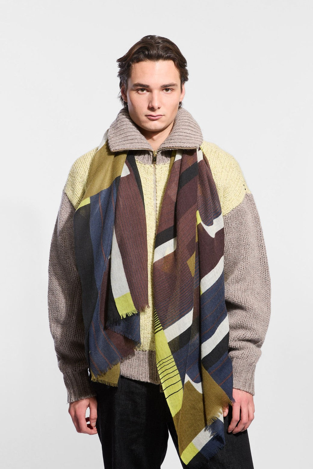 A male model wearing a chunky knit zip-up sweater with a high ribbed collar in beige and brown, layered with a large geometric color-blocked scarf in olive green, brown, navy, black, and cream.