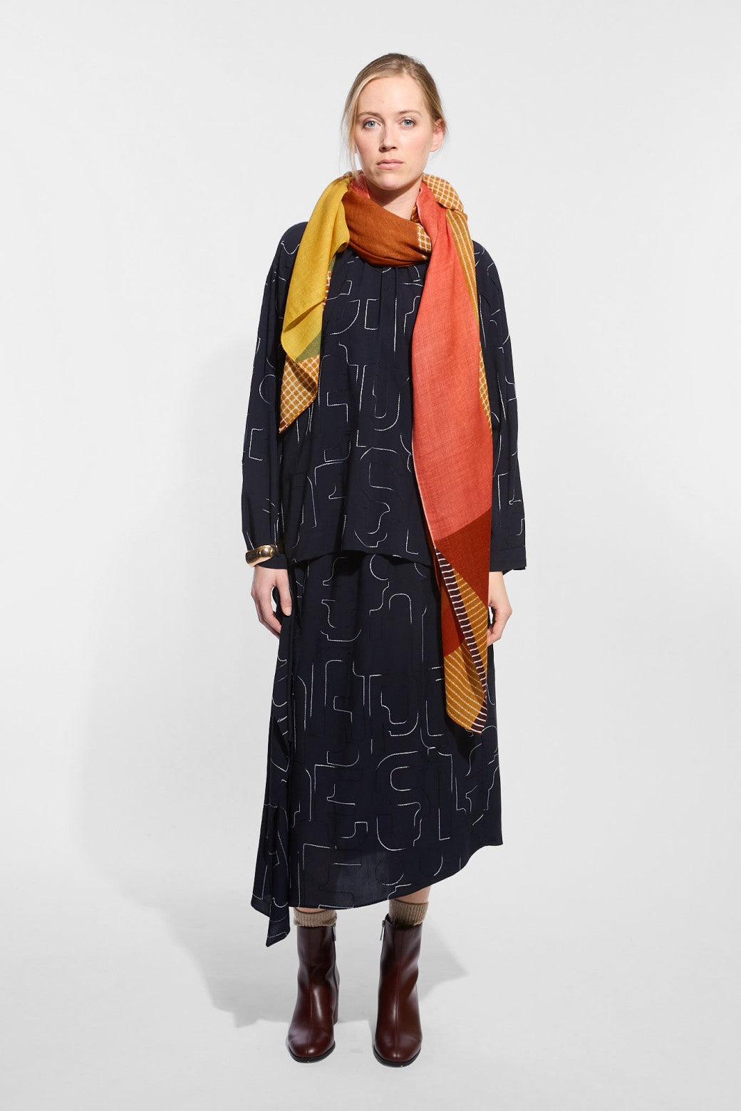 A model wearing a dark navy patterned dress, a mustard yellow and rust orange scarf, and dark brown ankle boots.