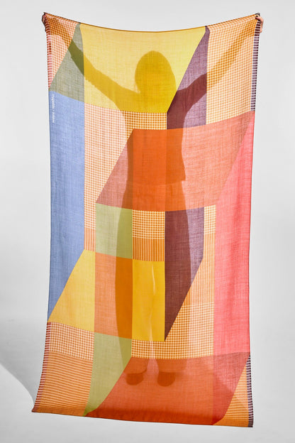 A lightweight rectangular scarf with a colorful geometric pattern, featuring a semi-transparent design with a printed silhouette of a figure with raised arms. The pattern includes blocks of orange, yellow, pink, blue, and green with some fine checkered details, shown hanging against a neutral light gray background.