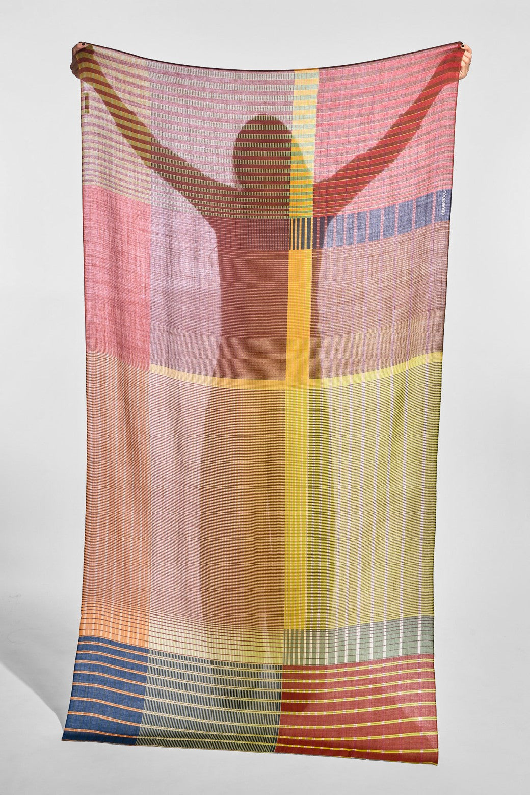 A lightweight striped scarf or shawl with a multicolor geometric pattern, held up by a person whose silhouette is cast onto the fabric against a plain light gray studio background.