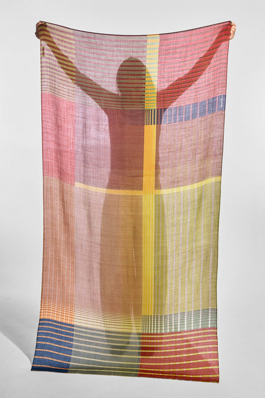 A lightweight striped scarf or shawl with a multicolor geometric pattern, held up by a person whose silhouette is cast onto the fabric against a plain light gray studio background.